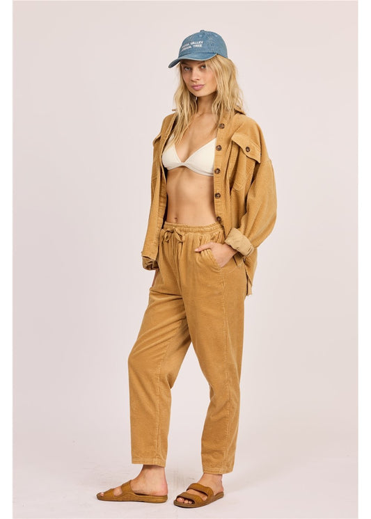 Woman wearing a beige jacket, pants, and cap with a white bikini underneath on a light pink background