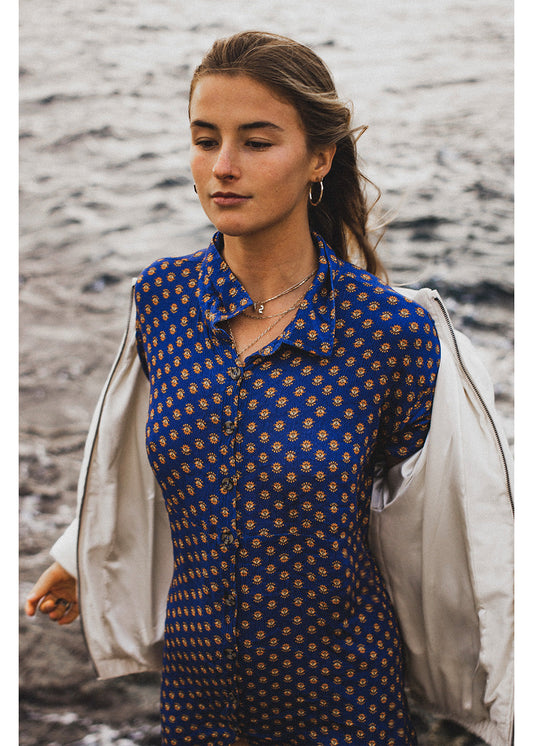 Woman wearing a blue patterned shirt with a beige jacket by a body of water.