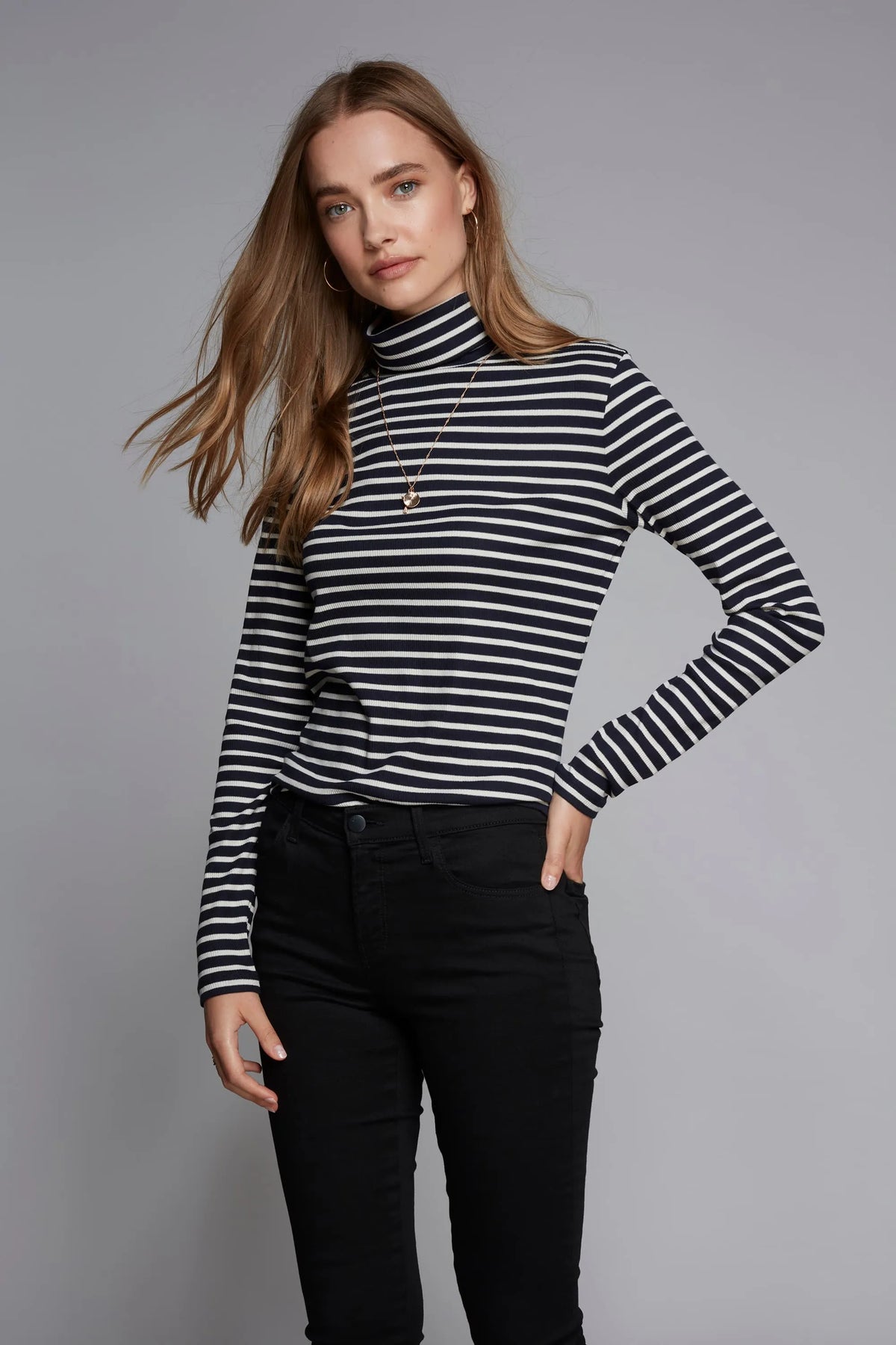 Woman wearing a black and white striped turtleneck and black pants on a gray background